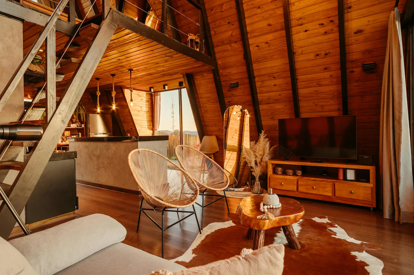 Interior of an A-frame cabin bedroom with a comfortable bed, wooden walls, and large windows overlooking nature.
